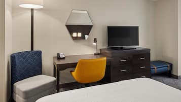 A hotel room with a bed, a desk with a computer, a chair, a lamp, a TV, and a small table.