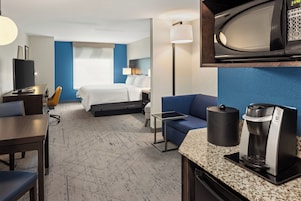 Quarto de hotel com cama, mesa, cadeira, máquina de café e micro-ondas.