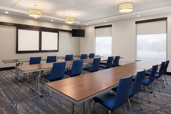 Un salón de conferencias con una larga mesa, sillas azules, un televisor de pantalla plana y ventanas amplias.