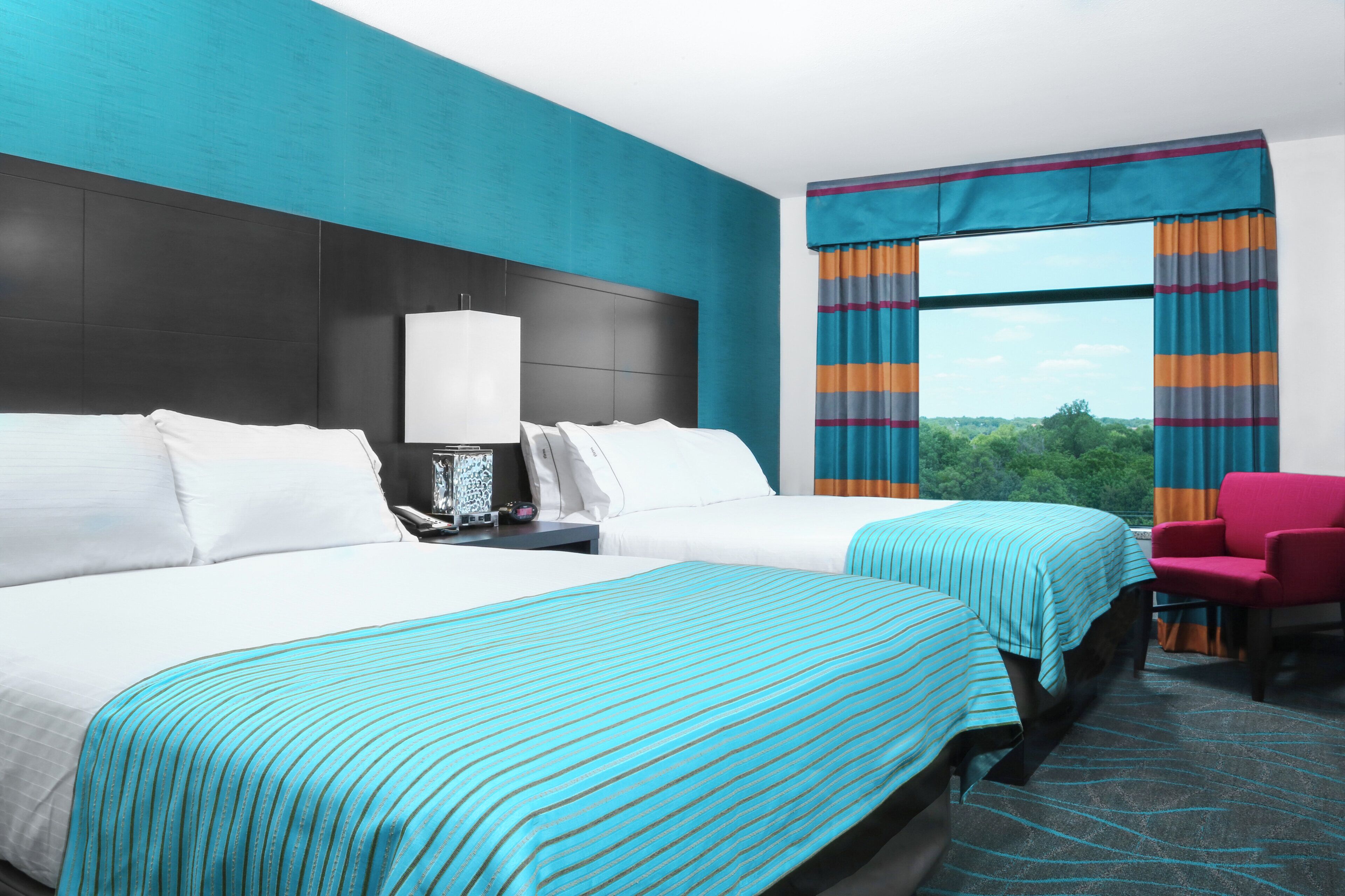 A hotel room with two beds, a nightstand, a lamp, a chair, and a window with curtains.