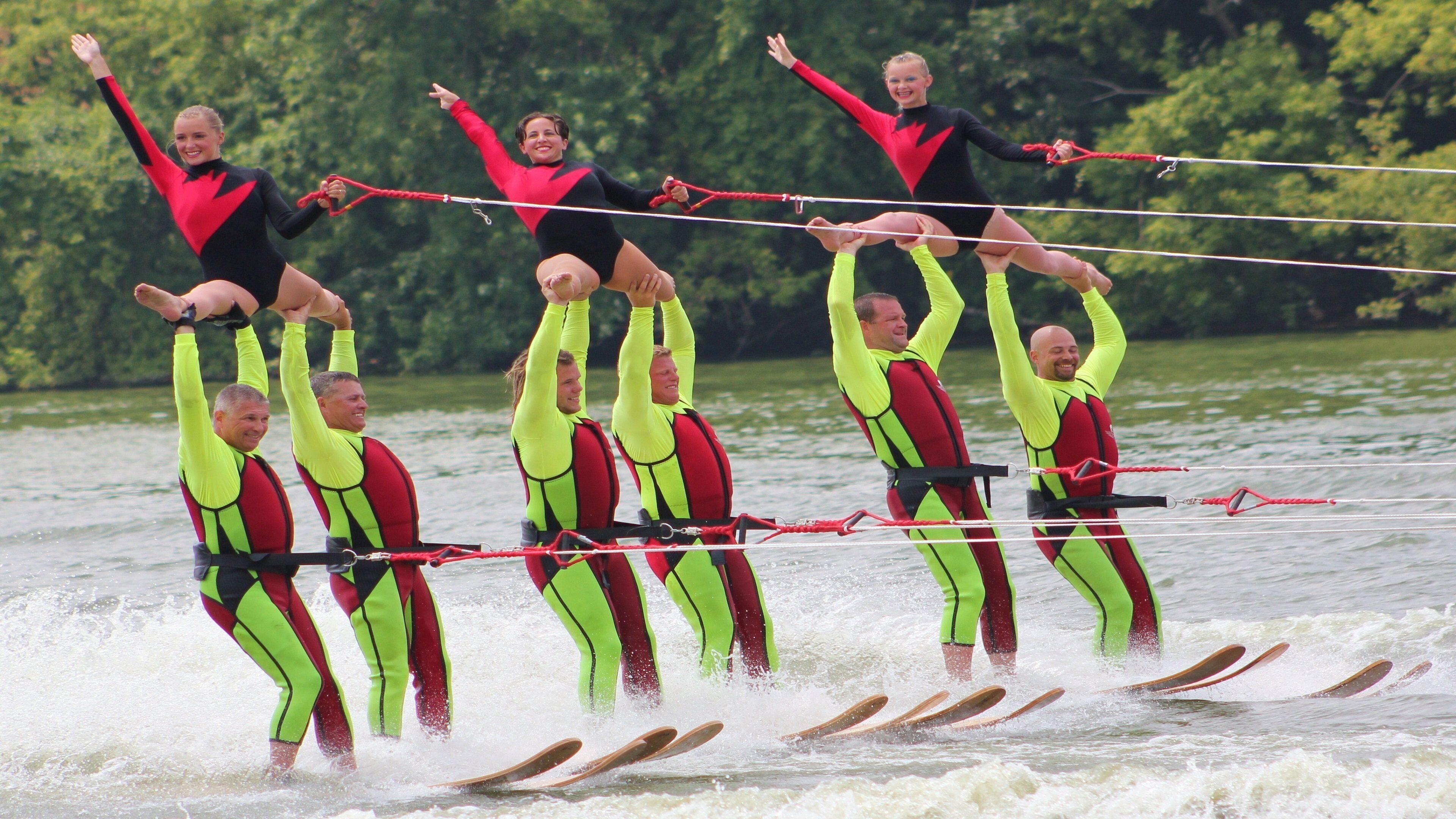 A group of people performing an acrobatic act on skis, with a body of water and trees in the background.