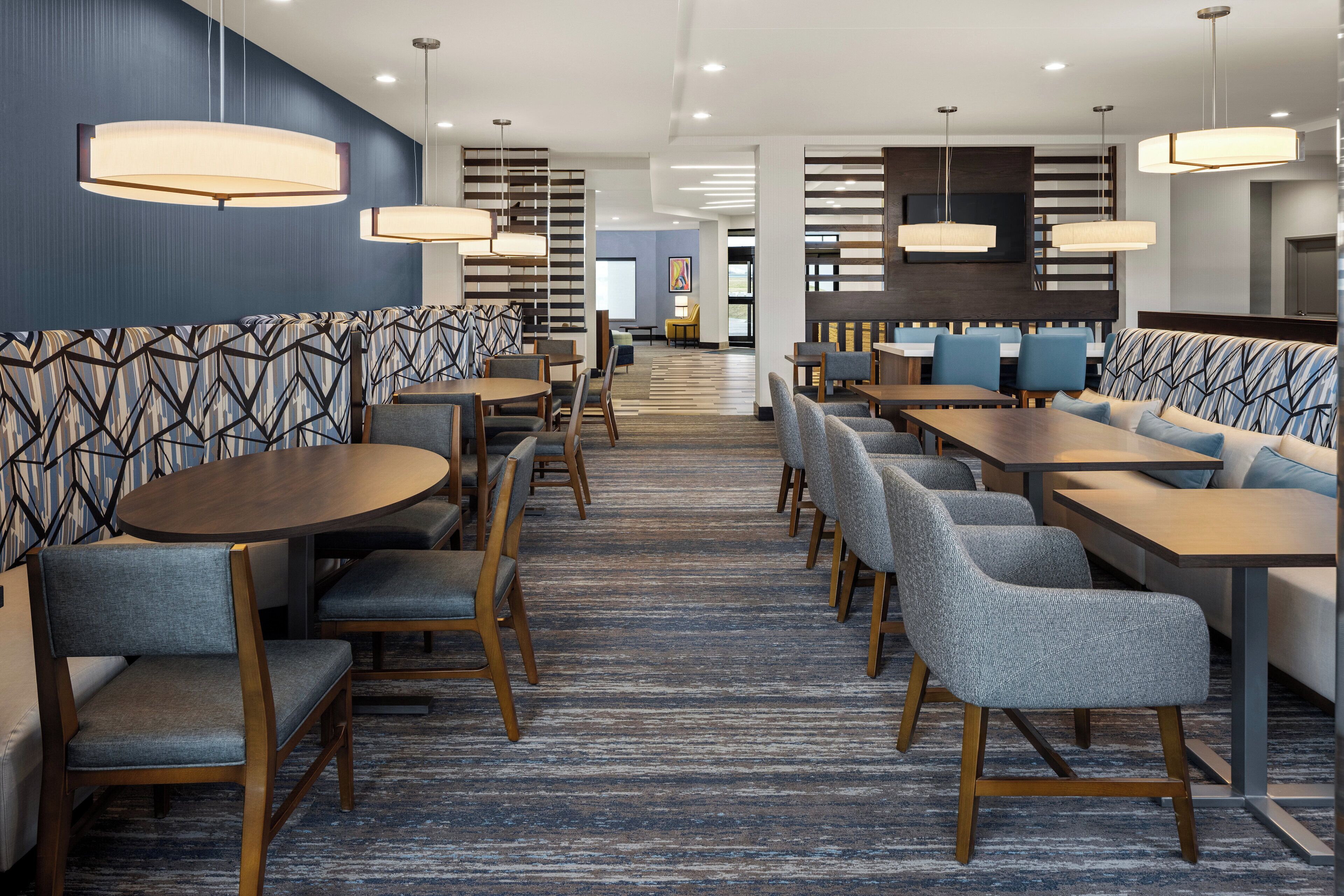 A modern hotel lobby with a central seating area, multiple tables, and contemporary lighting fixtures.
