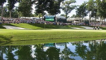 Un campo de golf con una multitud de espectadores, un estanque que refleja la escena y un marcador en el fondo.