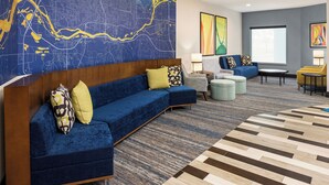 A modern hotel lobby with a large map mural, a blue sofa with yellow cushions, a gray armchair, and a patterned rug.