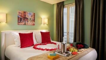 Habitación de hotel con cama, almohadas rojas, un cuadro en la pared, ventana con cortinas y una bandeja con champán y flores.