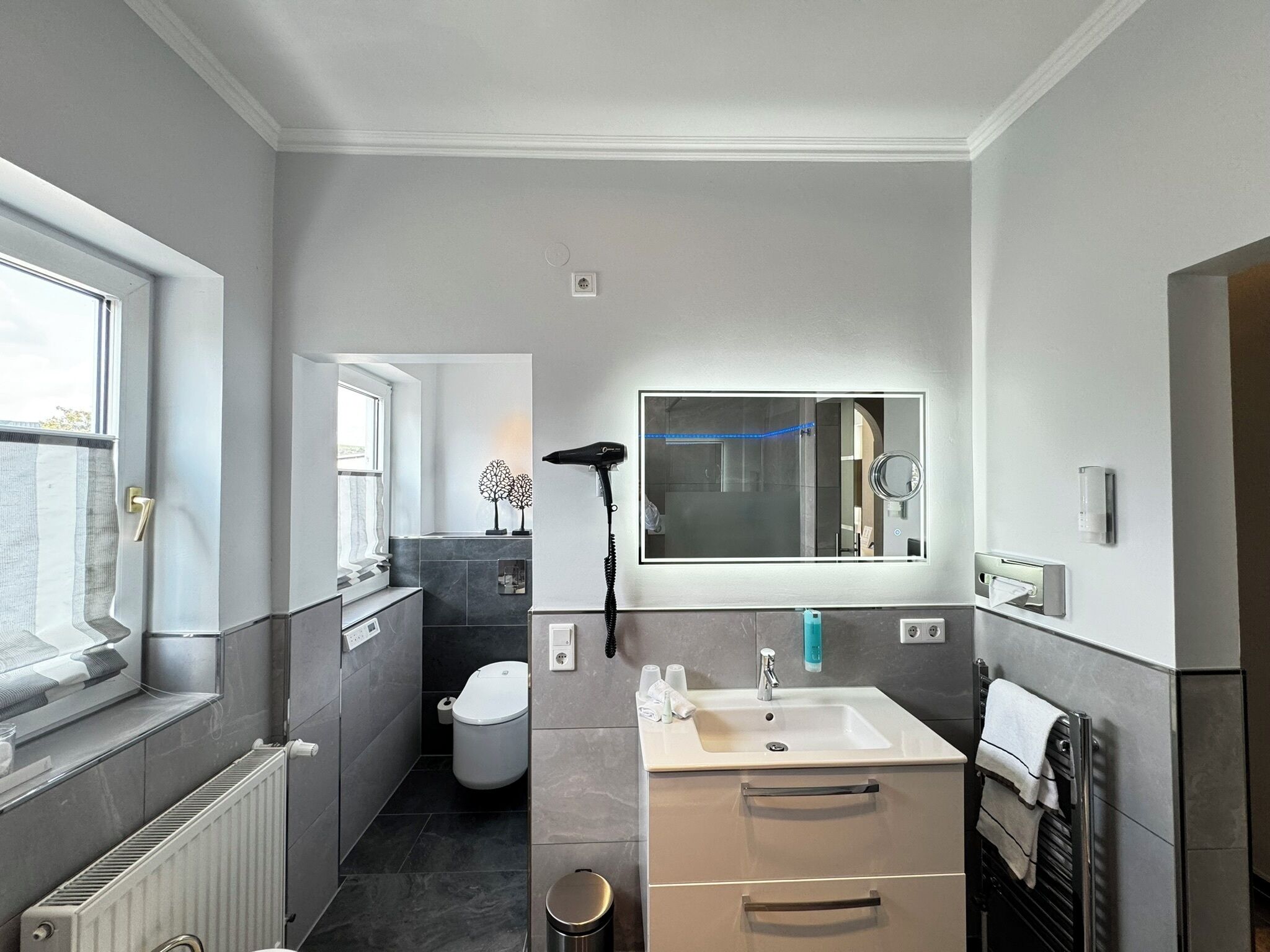 Un baño moderno con un espejo grande, un lavabo y un inodoro. Hay una ventana que proporciona luz natural y un radiador a la izquierda.
