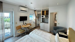 Habitación de hotel con cama, escritorio con silla, pequeña mesa de comedor con sillas, kitchenette y televisor montado en la pared.
