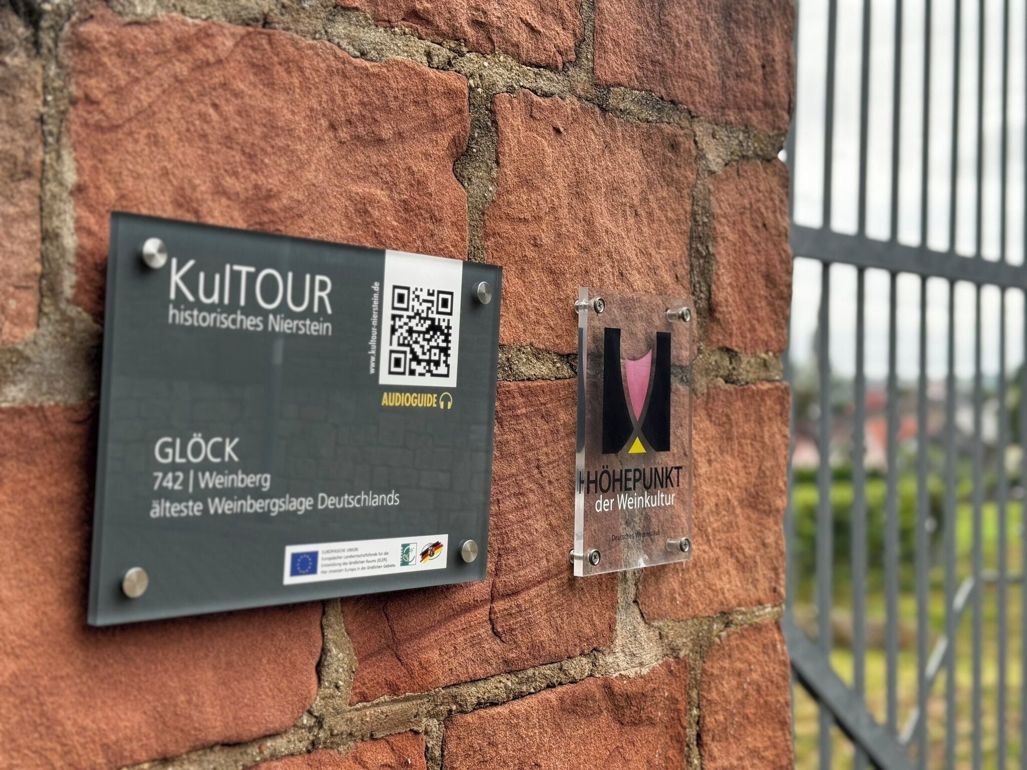 A brick wall with two plaques. One plaque is labeled 'KulTour historisches Nierstein' and includes a QR code. The other plaque is labeled 'HÖHEPUNKT der WeinKultur' with a logo.