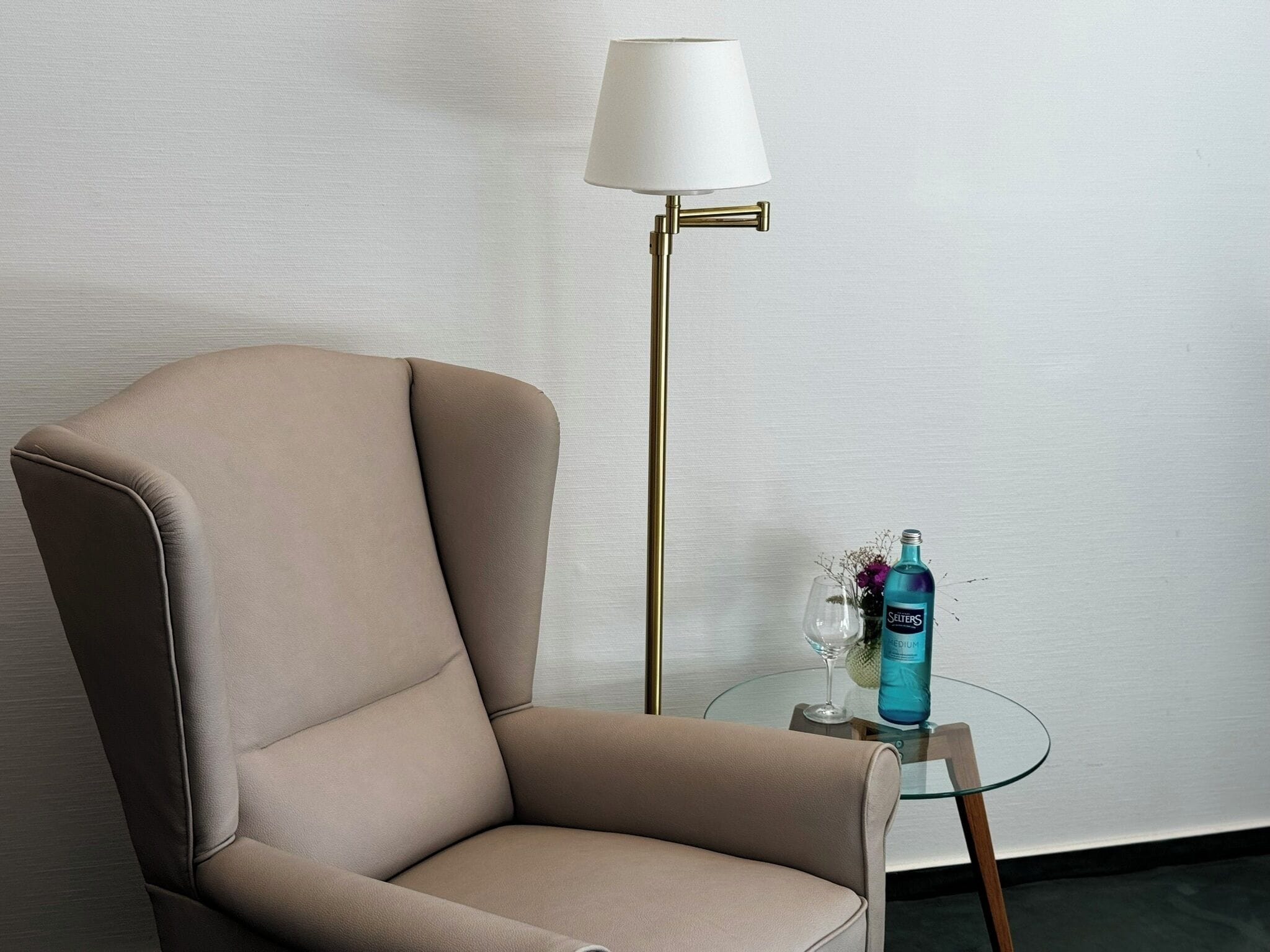 Un fauteuil beige, une lampe sur pied et une table en verre avec une bouteille et un verre.