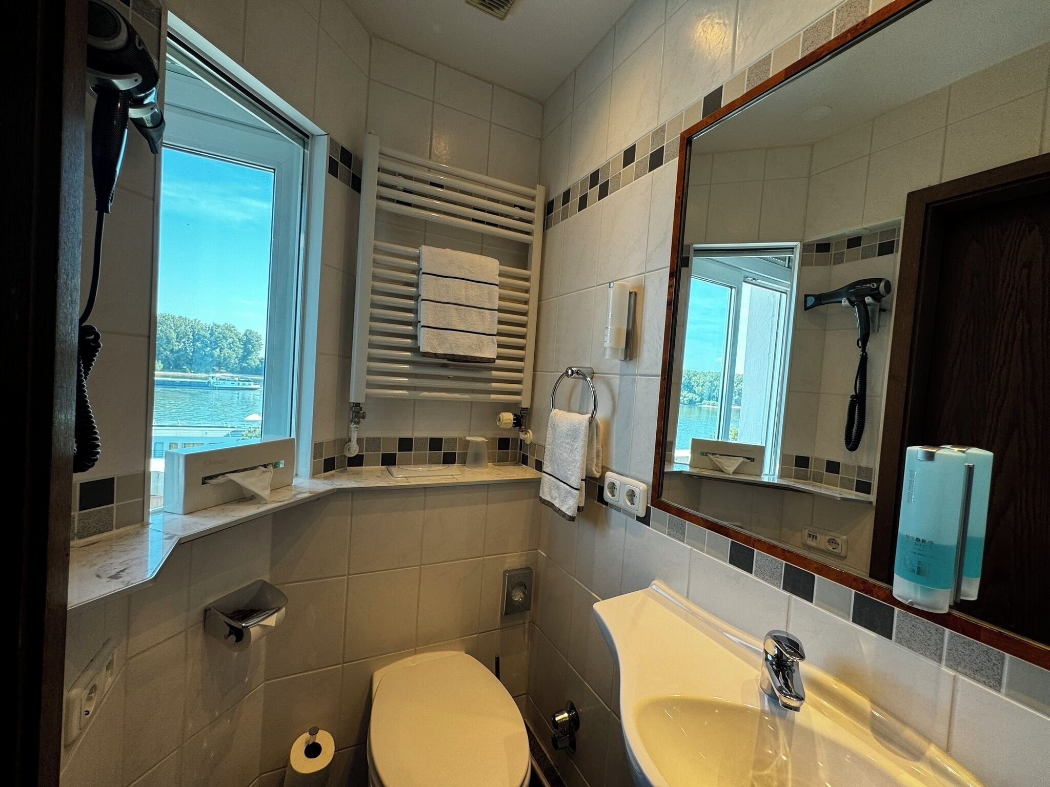 A bathroom with a toilet, sink, towel rack, and a window with a view of a body of water.