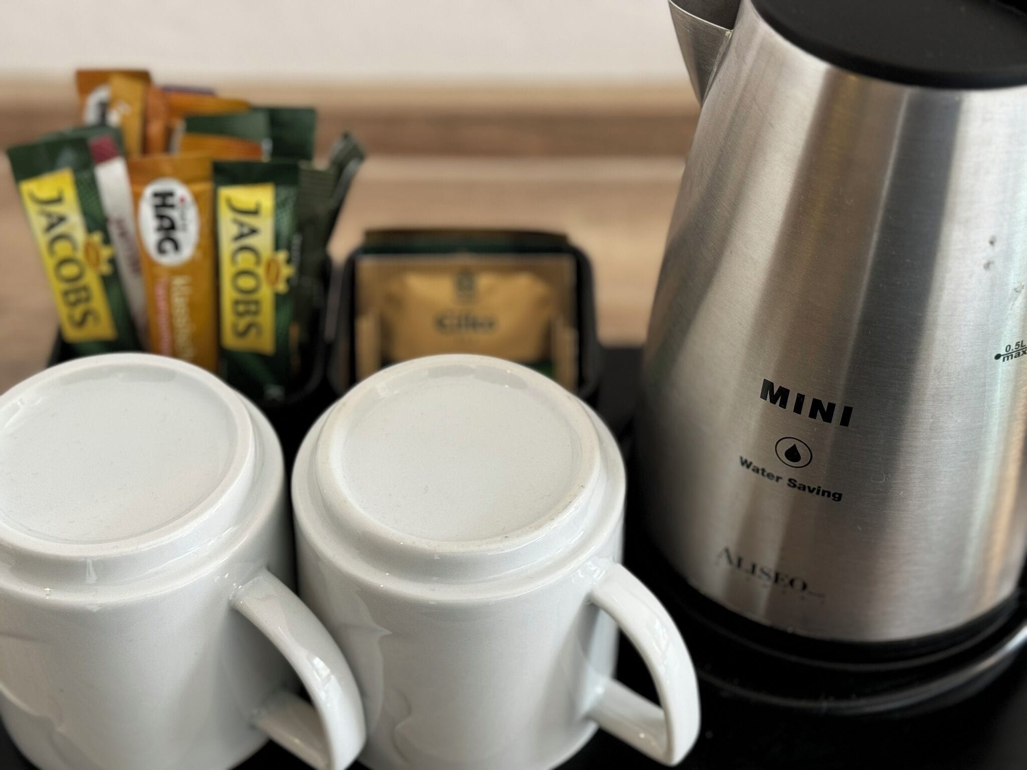 Deux tasses à café blanches, une cafetière en acier inoxydable portant l’inscription « MINI, pour une consommation d’eau réduite », et plusieurs paquets de café portant l’inscription « JACOBS ».