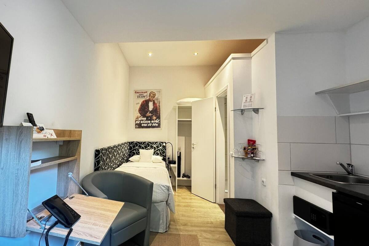 A compact hotel room with a bed, a desk with a telephone, a small kitchenette, and a poster on the wall.