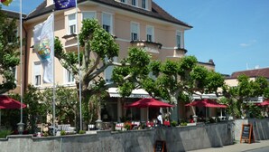 Un hotel con terraza al aire libre, un asta con la bandera alemana y un cartel.