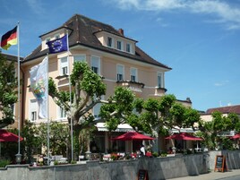 A hotel with outdoor seating, a flagpole with the German flag, and a signboard.