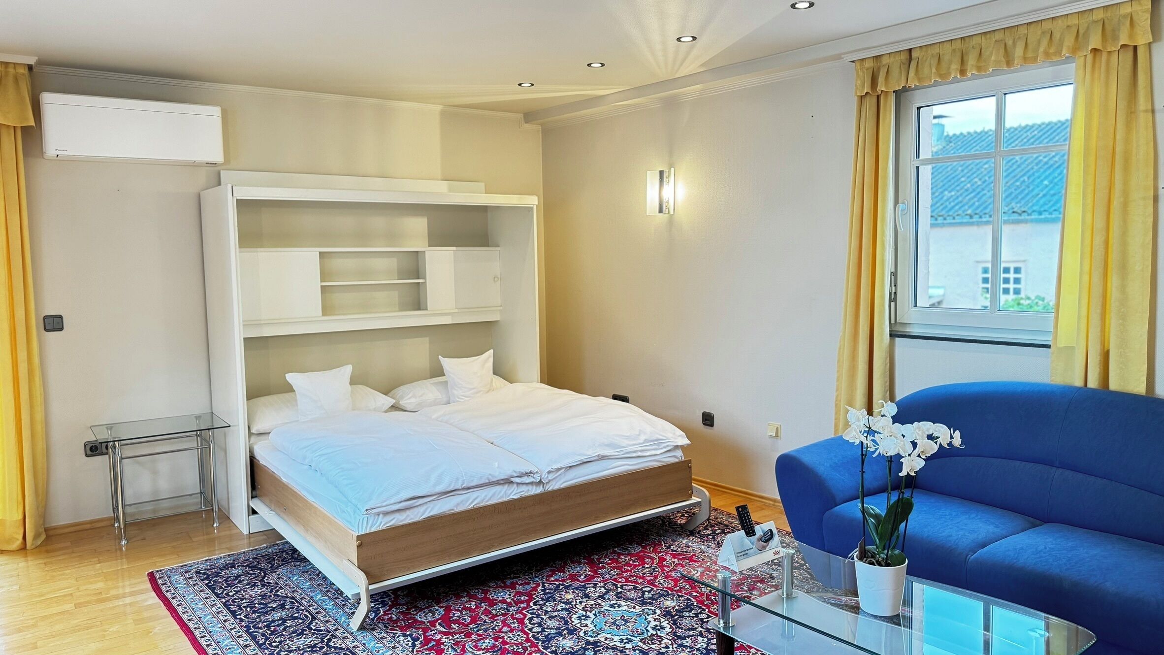 Un dormitorio moderno con cama, estanterías empotradas, una mesa de centro de vidrio, un sofá azul y una ventana con cortinas.