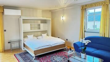 Un dormitorio moderno con cama, estanterías empotradas, una mesa de centro de vidrio, un sofá azul y una ventana con cortinas.