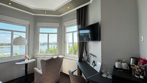 Habitación de hotel con un gran ventanal que ofrece vistas a un lago, un escritorio con televisión de pantalla plana, una silla y una mesita con un florero.
