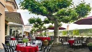 Un café al aire libre con mesas y sillas, un árbol grande y vistas al río.