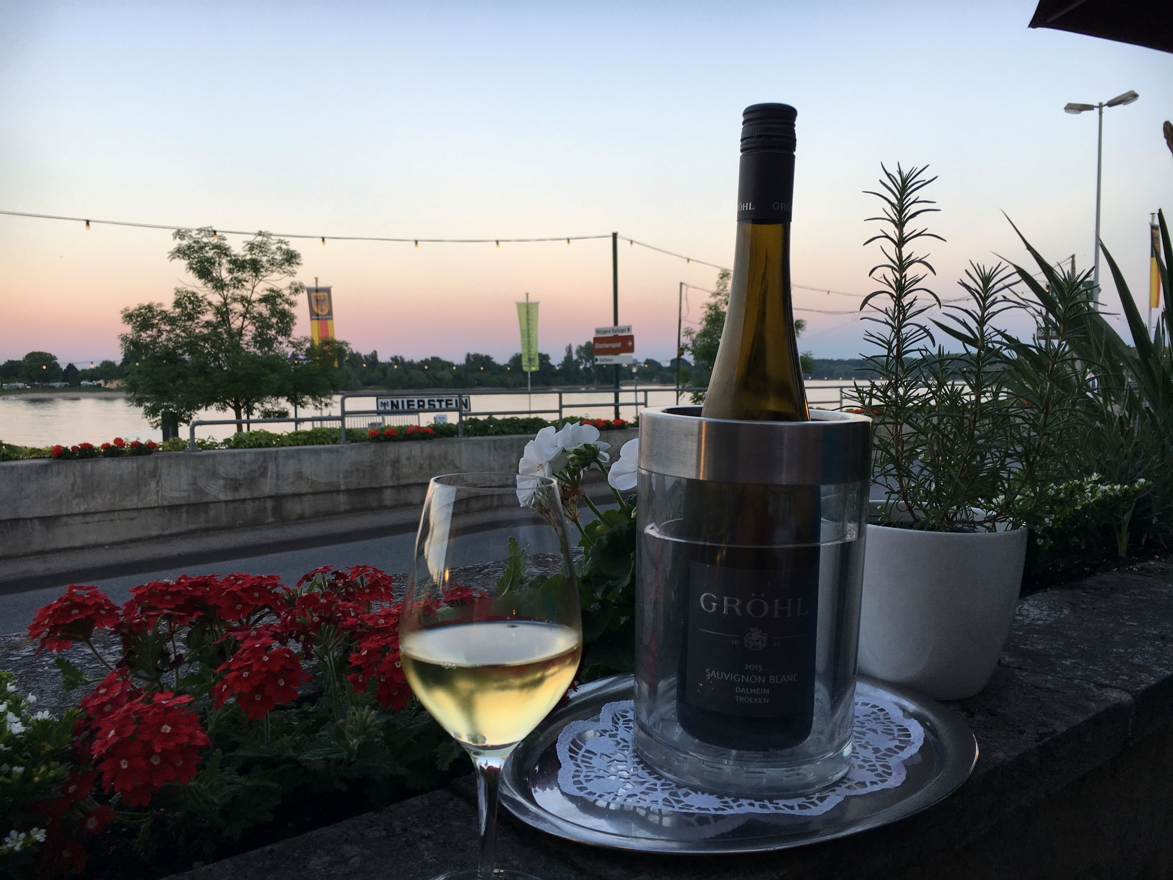Una botella de vino, una copa de vino y una planta en maceta sobre una mesa, con vistas a un río y un puente al fondo.