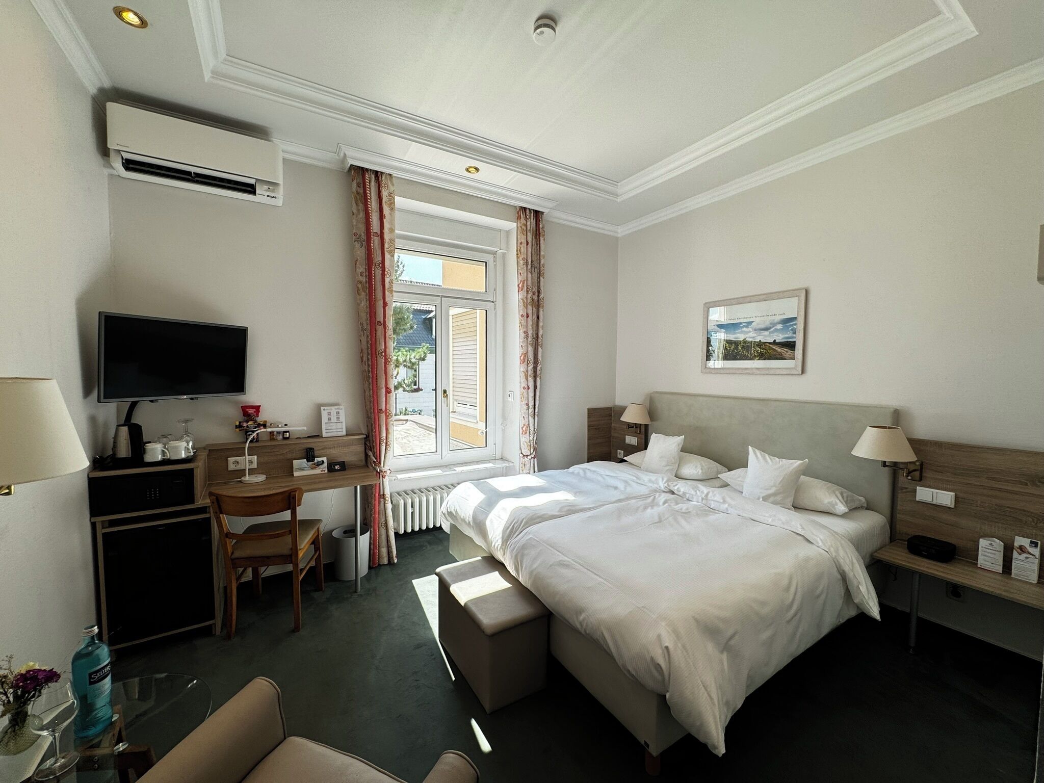 Quarto de hotel com cama, mesa de cabeceira, luminária, escrivaninha com computador e televisão.