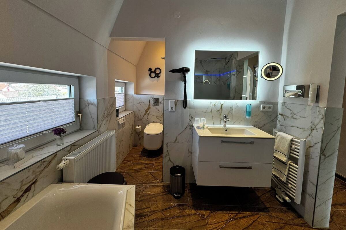 A modern bathroom with a bathtub, sink, and toilet. There is a window with blinds, a towel rack, and a mirror above the sink.