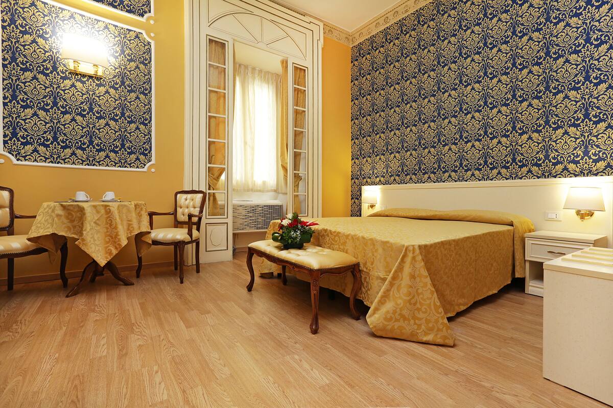 A hotel room with a bed, a bedside table, a chair, a small table with a tablecloth, and a window with curtains.