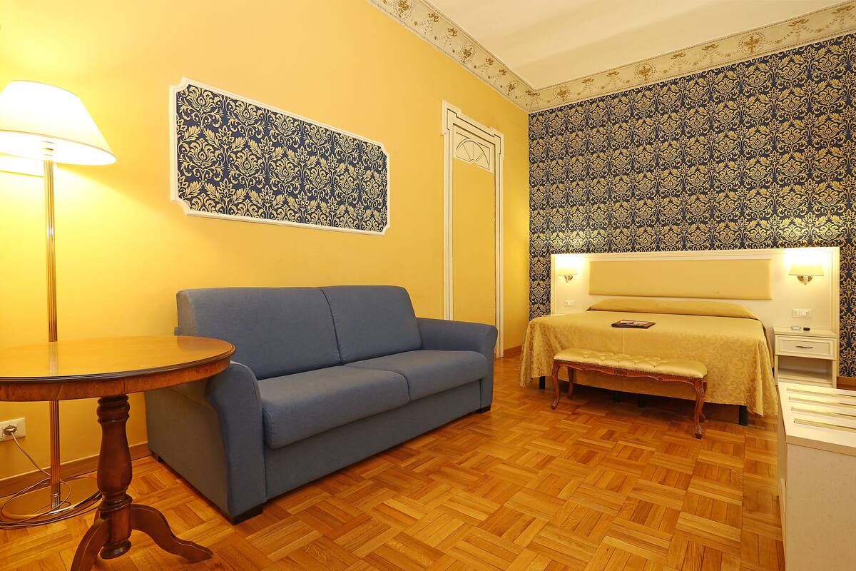 A room with a blue sofa, a round wooden table, a bed with a white headboard, and patterned wallpaper.