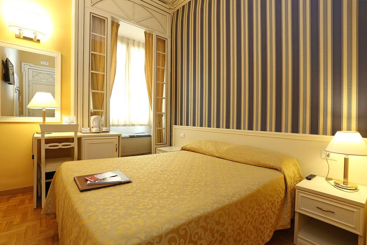 A hotel room with a bed, a desk, a chair, a nightstand, a lamp, and a mirror.