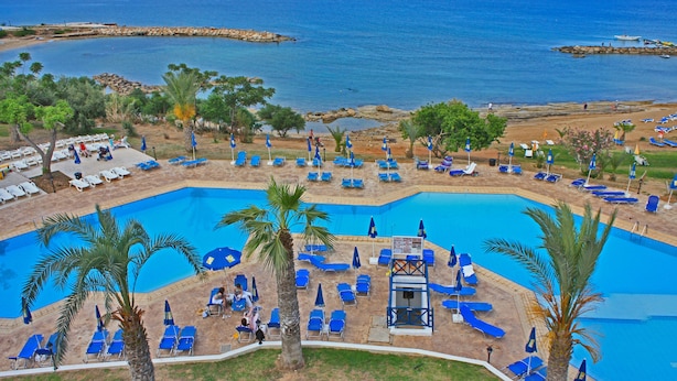 A resort pool area with multiple sun loungers and palm trees, surrounded by a clear view of the sea. - Myroandrou Beach Apartments (Paralimni)
