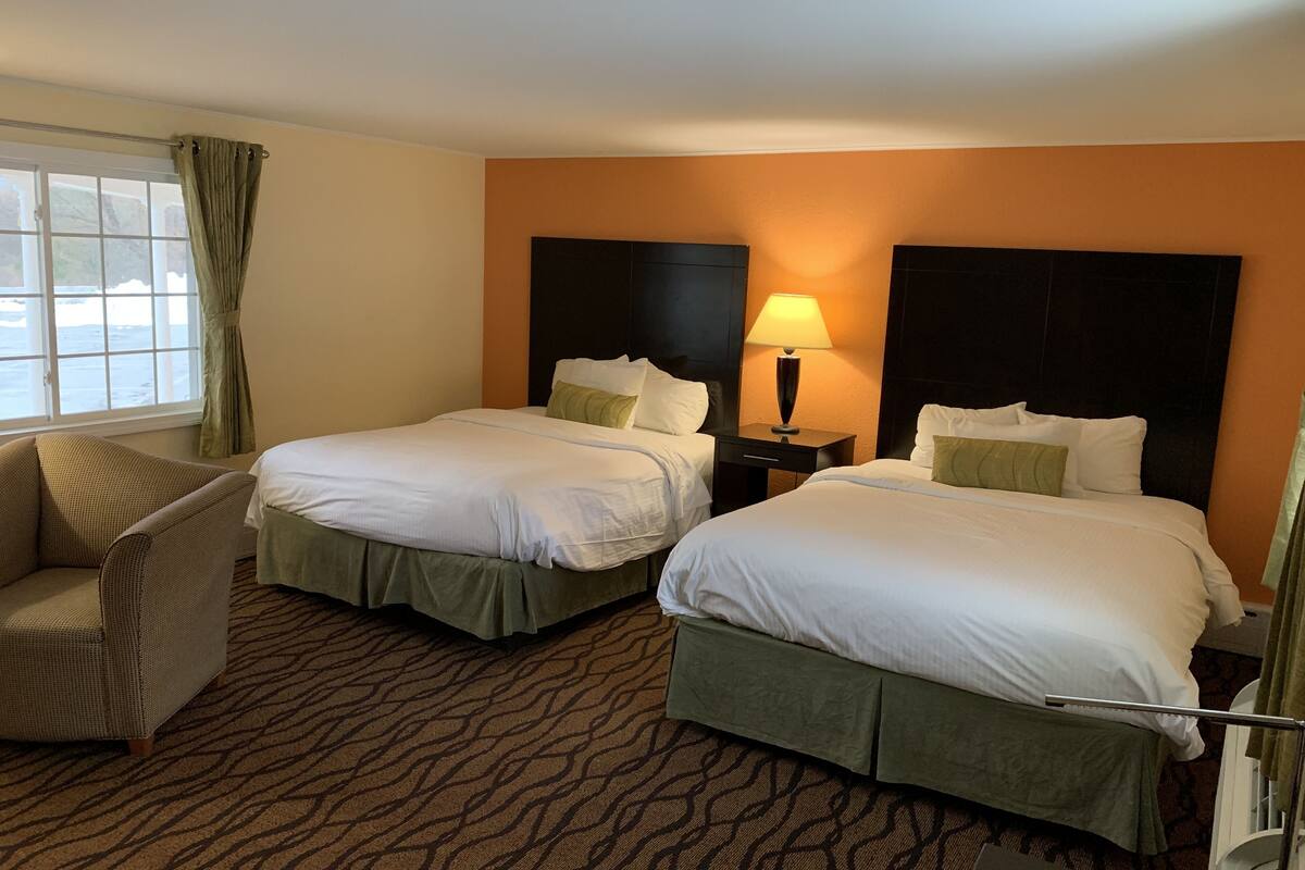 A hotel room with two beds, a chair, a nightstand with a lamp, and a window with curtains.