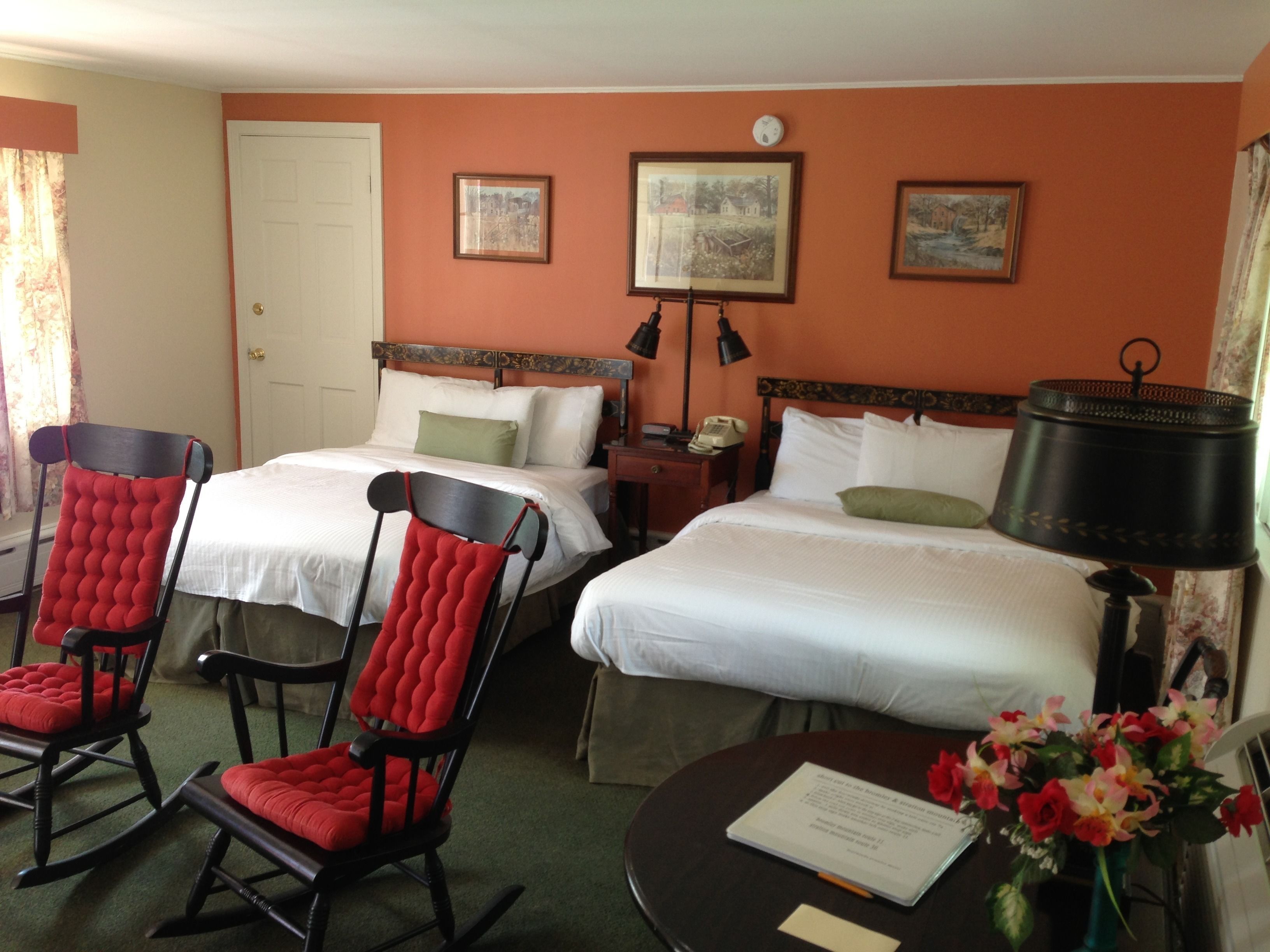 A hotel room with two beds, a desk, a chair, a lamp, and a vase with flowers.