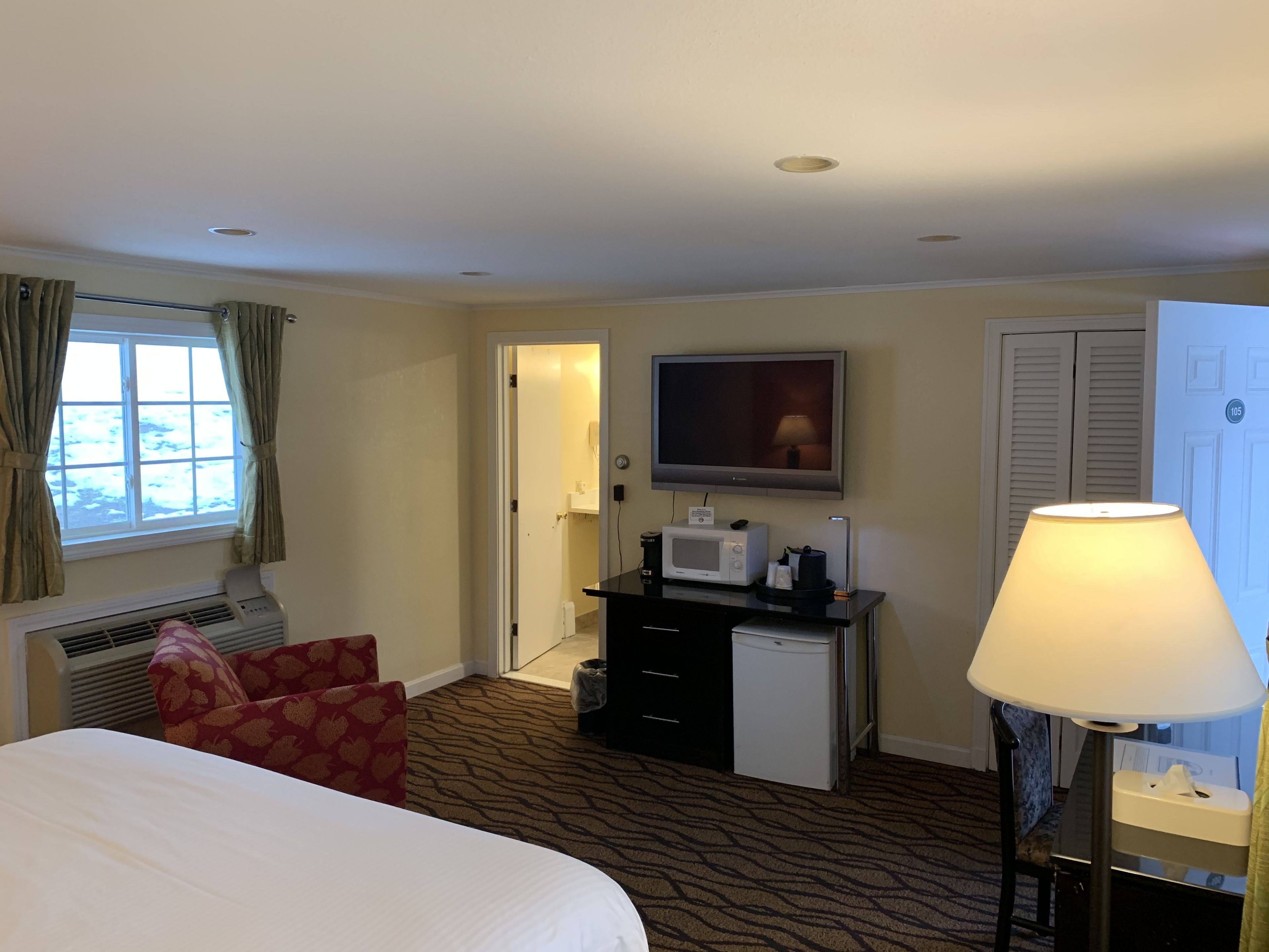 Habitación de hotel con cama, escritorio con televisor, lámpara, microondas y refrigerador pequeño.