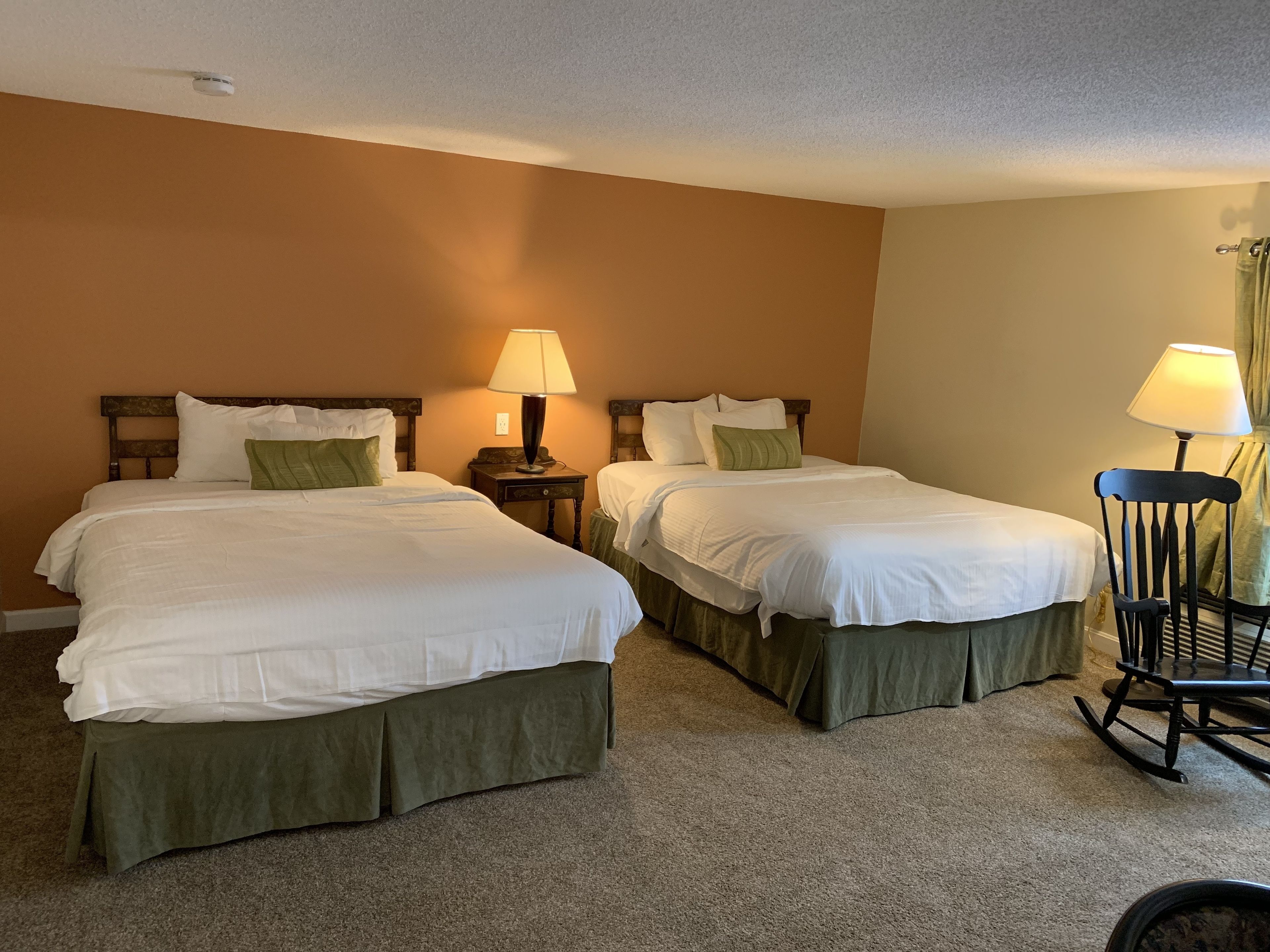 A hotel room with two beds, each with a green skirt, a nightstand with a lamp, and a rocking chair.