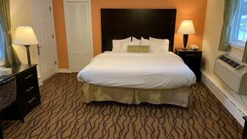 A hotel room with a large bed, a dresser, a nightstand with a lamp, a telephone, and an air conditioning unit.