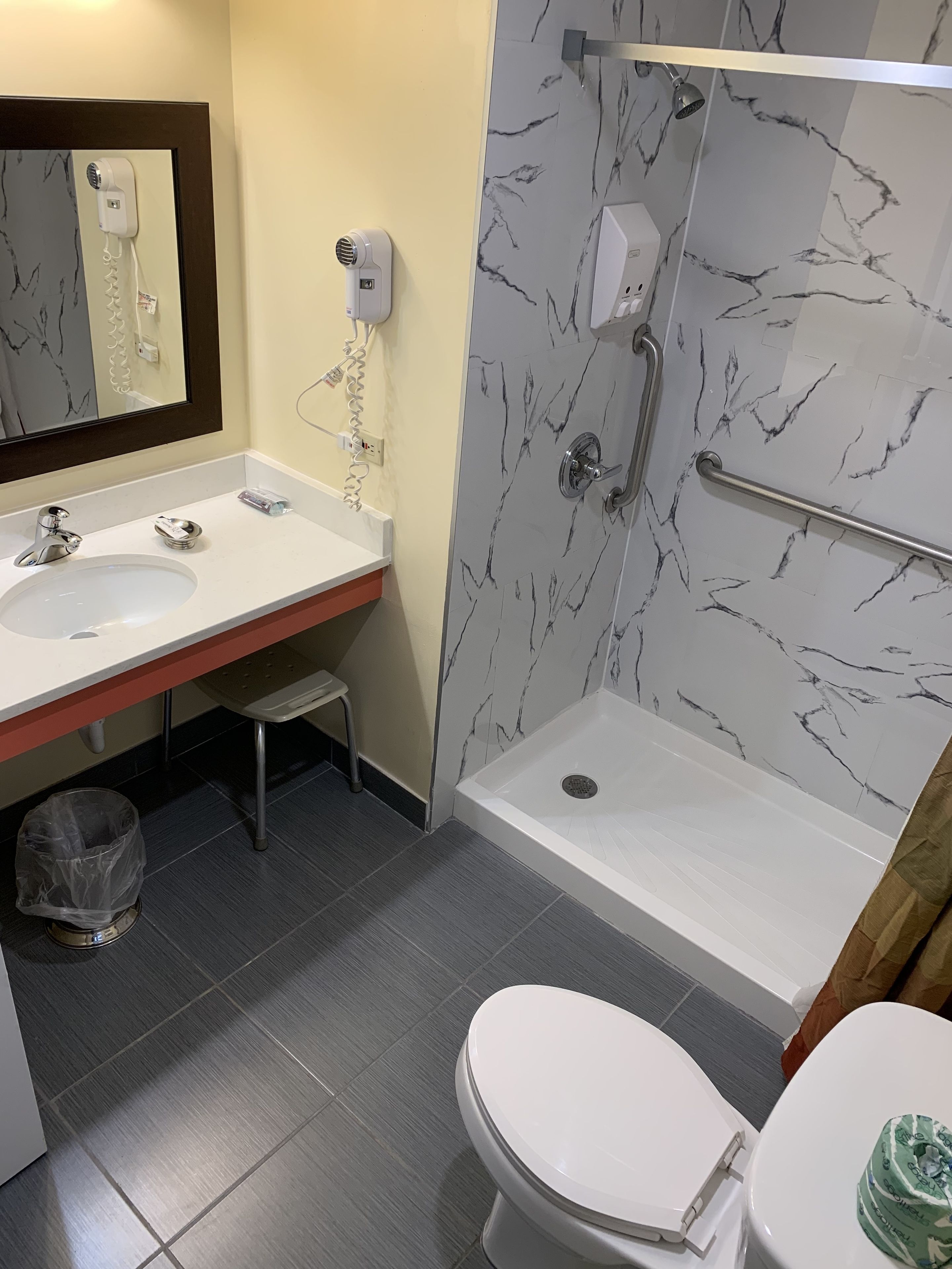 A bathroom with a white sink, mirror, and toilet, a shower with a marble wall, and a trash can.