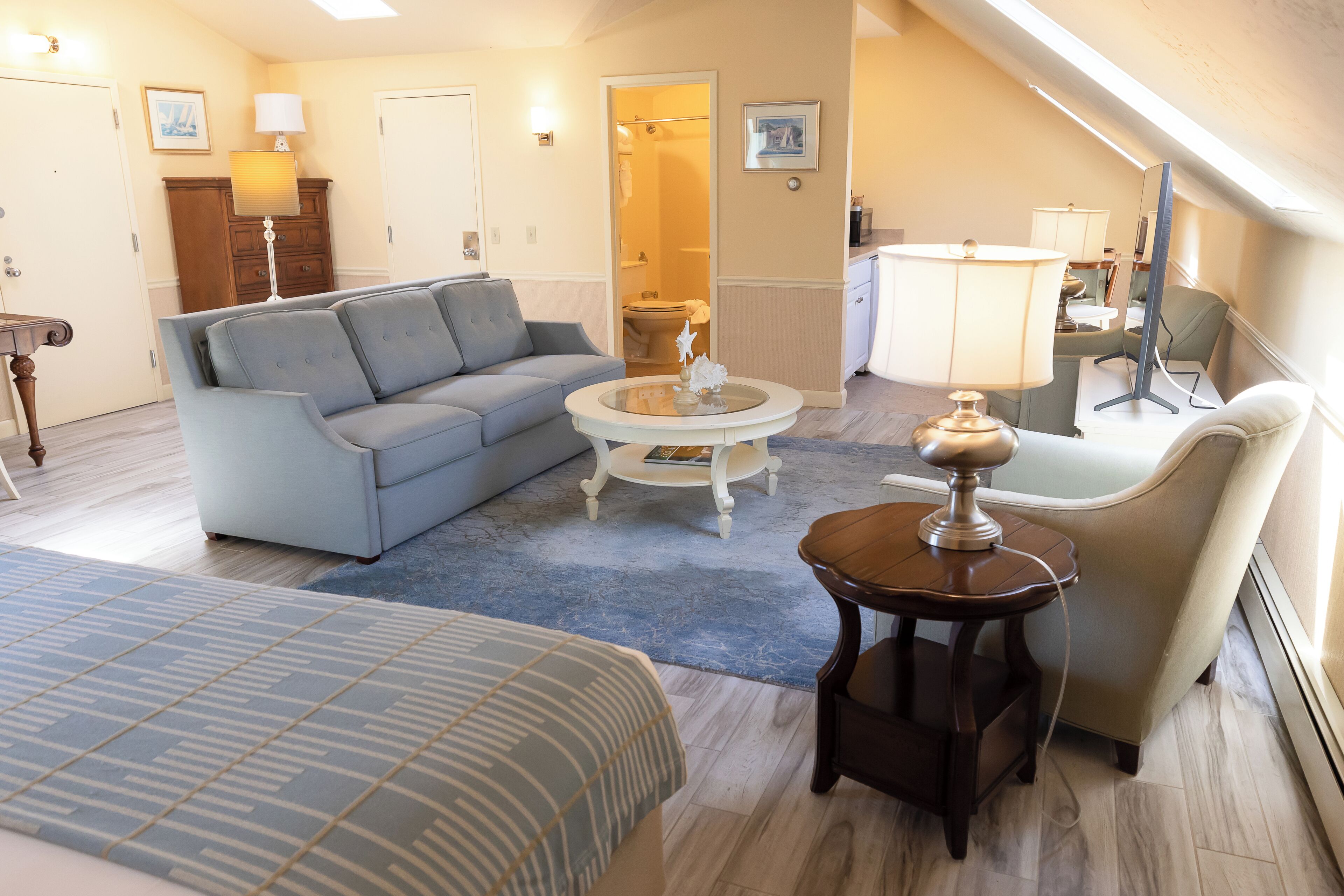 A well-lit living room with a sofa, a coffee table, a side table, and a chair. There is a door leading to a bathroom.