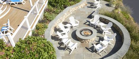 Un espacio de estar al aire libre circular con sillas Adirondack blancas y un pozo de fuego de piedra, rodeado por un muro de piedra y una terraza de madera con sillones.