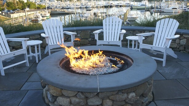 A stone fire pit with four white Adirondack chairs around it, overlooking a marina with boats. - Anchor In Distinctive Waterfront Lodging (Hyannis)