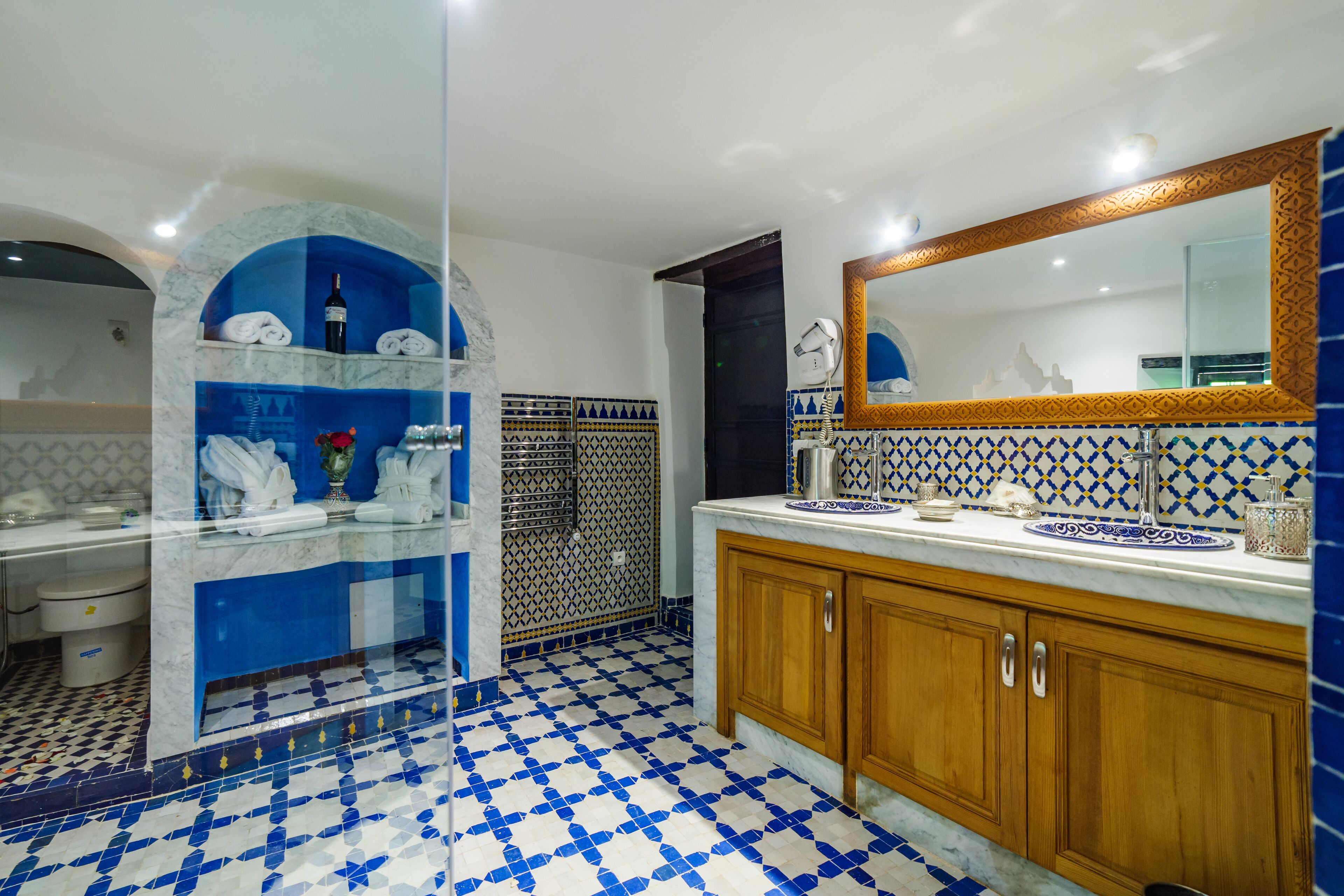 Une salle de bain avec un sol à carreaux bleus et blancs, un lavabo en marbre, des meubles en bois et un grand miroir doté d’un cadre décoratif.