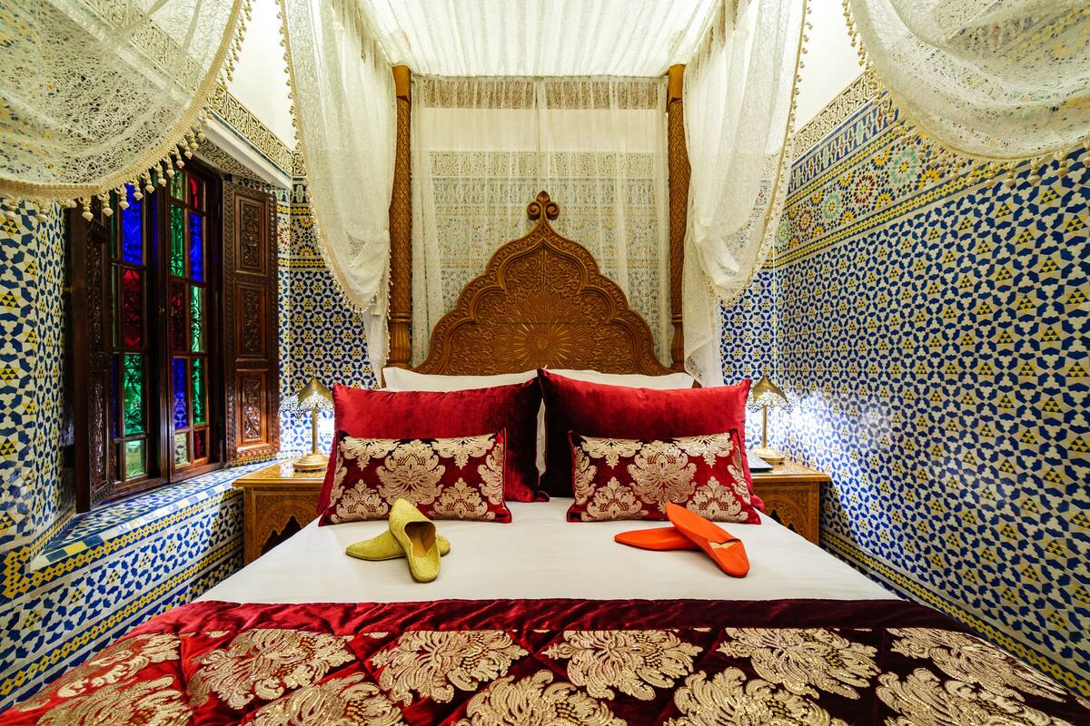 A room with a large bed, ornate headboard, patterned walls, and a window with stained glass.