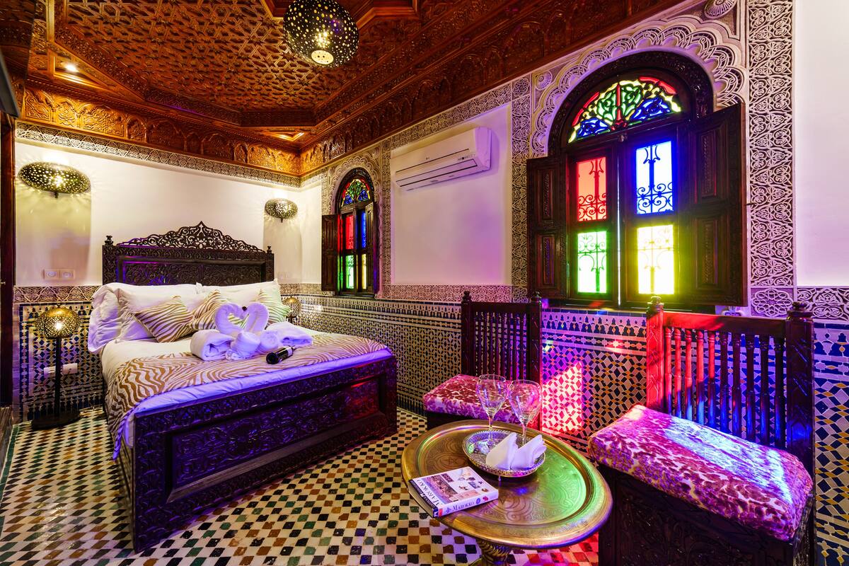 A room with a checkered floor, a bed with ornate headboard, a bench, and a table with a book and glasses.