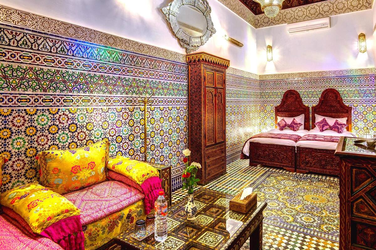 A room with intricate tile work, a bed with a wooden headboard, a sofa with colorful cushions, and a chandelier.
