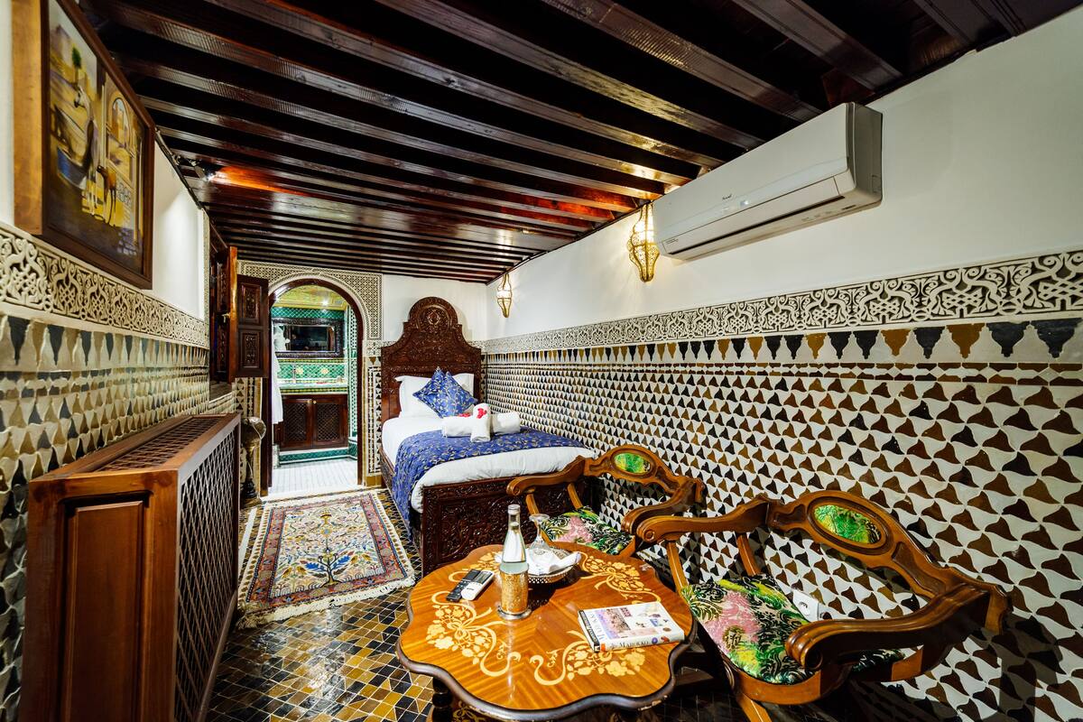 A traditional Moroccan room with a patterned tile wall, a wooden ceiling, a bed with a blue and white bedspread, and a small table with a bottle and a book.