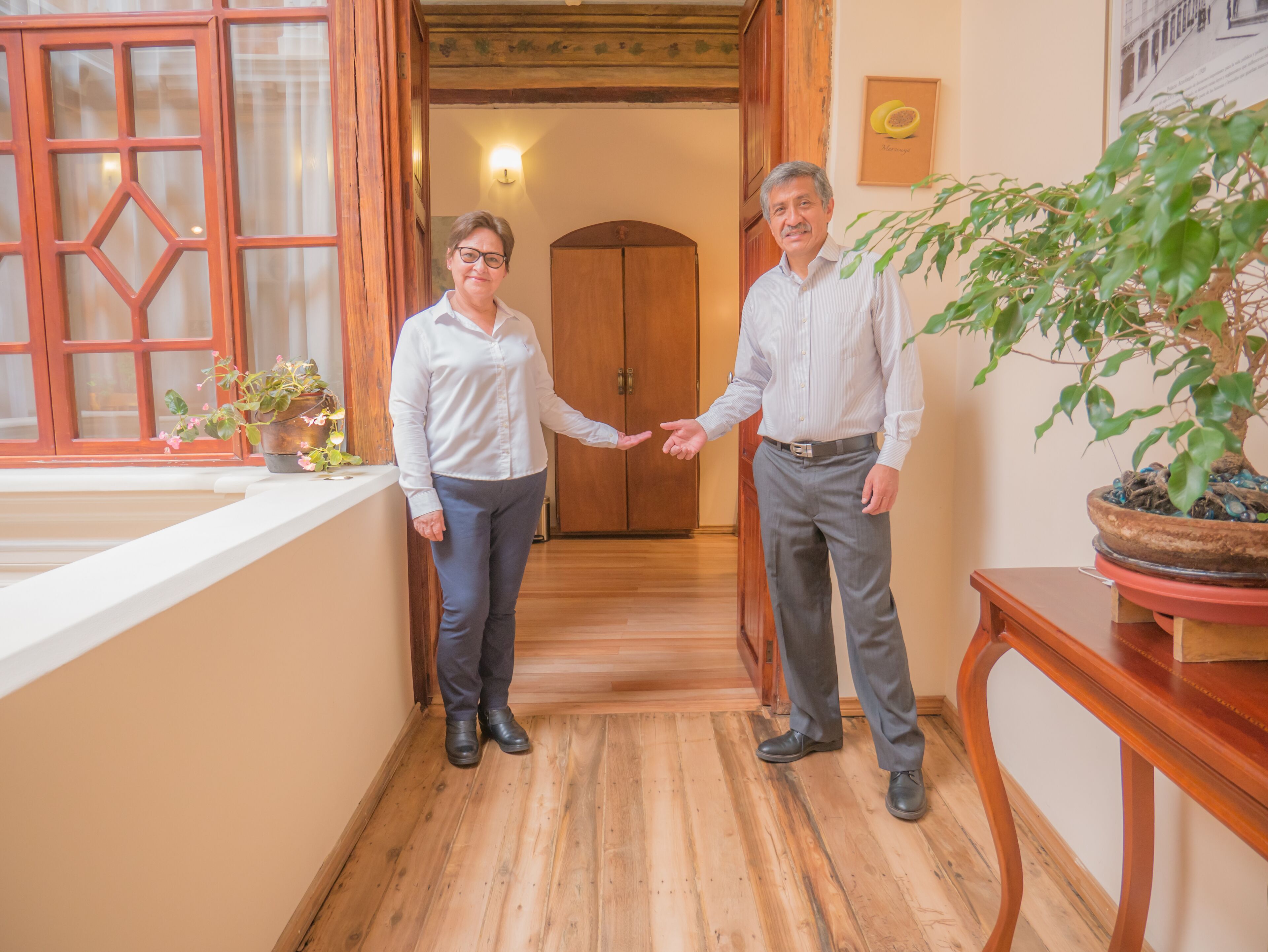 Dos personas estrechan la mano en un pasillo con suelo de madera, una puerta de madera y una planta en maceta sobre una mesa.