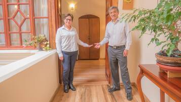 Dos personas estrechan la mano en un pasillo con suelo de madera, una puerta de madera y una planta en maceta sobre una mesa.