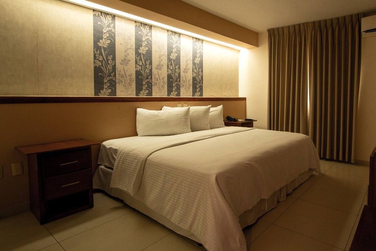 A hotel room with a large bed, a bedside table with a lamp, a wooden nightstand, and a mirror on the wall.