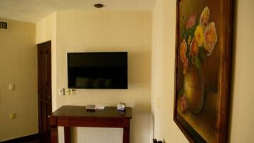 A room with a flat-screen TV mounted on the wall, a wooden table with a remote control, and a painting of flowers on the wall.