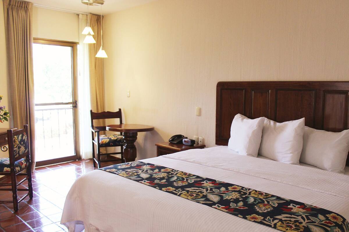 A hotel room with a large bed, a wooden headboard, a round table with a chair, a telephone, and a window with curtains.