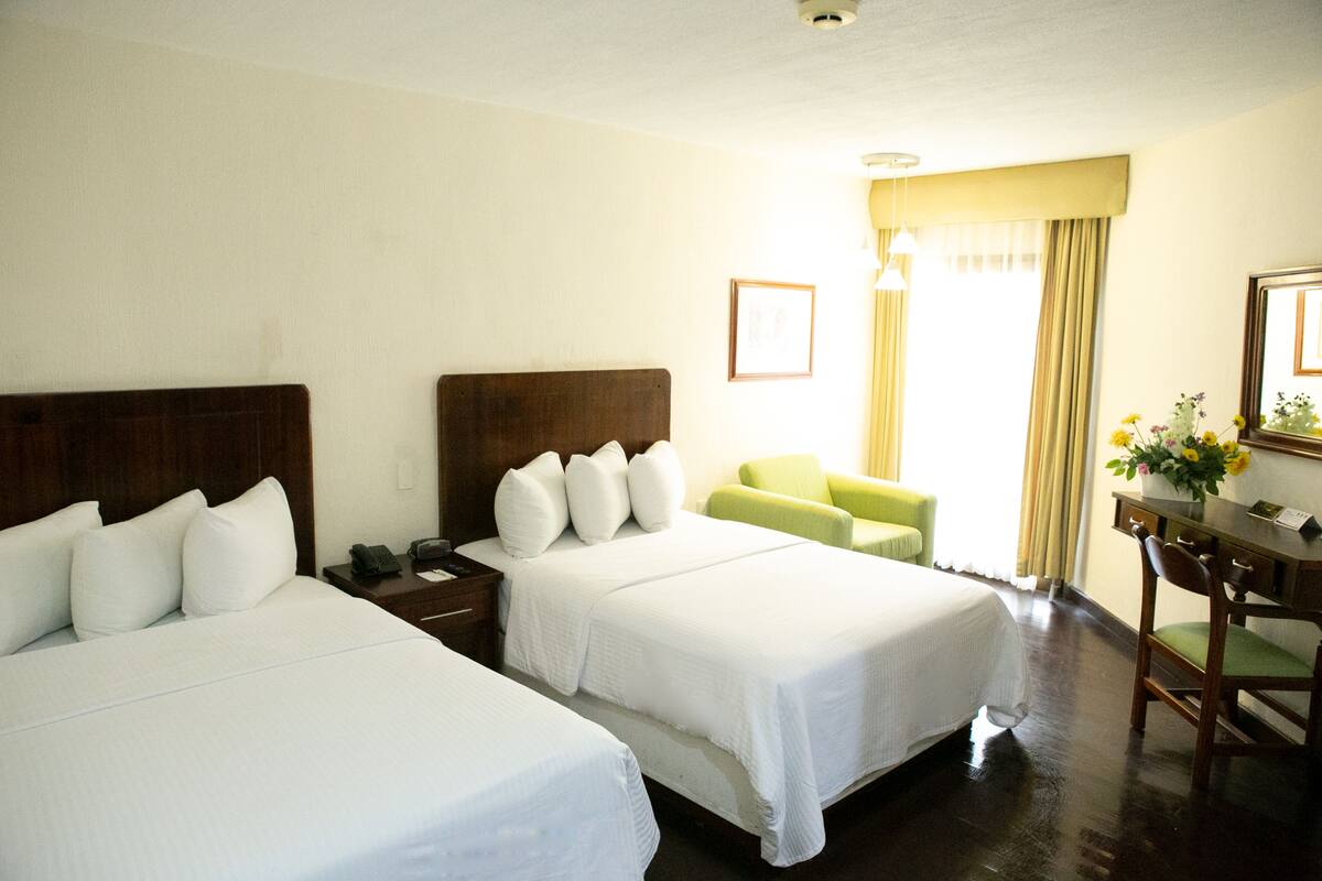 A hotel room with two beds, a desk, a chair, and a window with curtains.