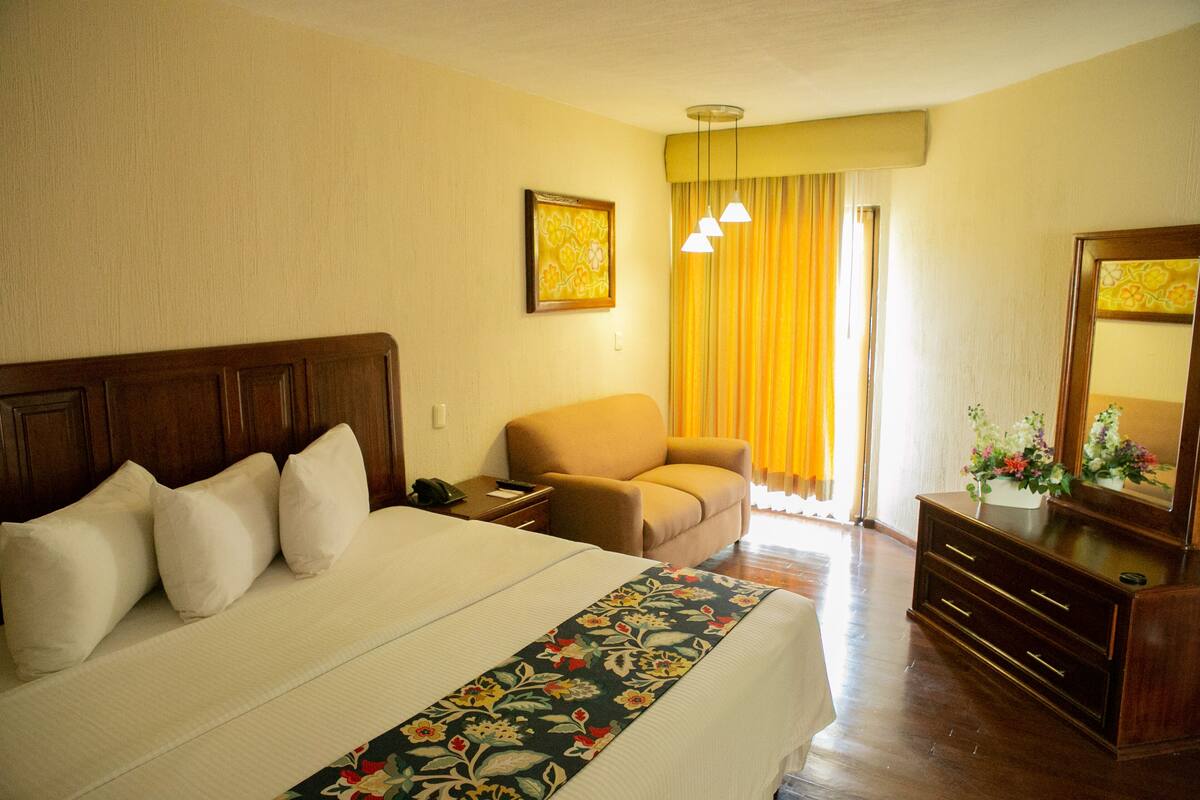 A hotel room with a large bed, a wooden nightstand, a chair, a dresser, and a mirror.